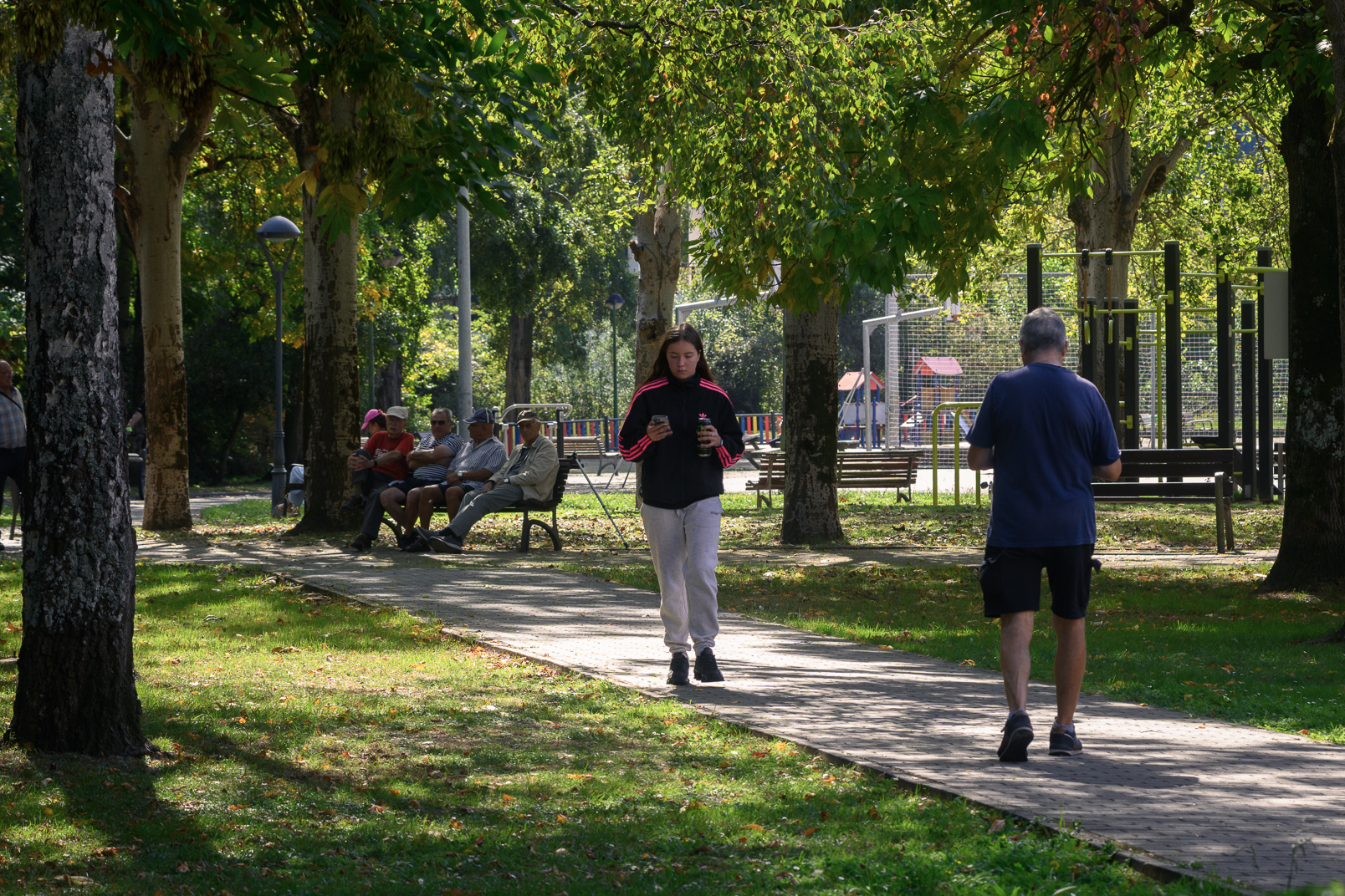 Personas pasean y descansan en los bancos de un parque arbolado de Llaranes.